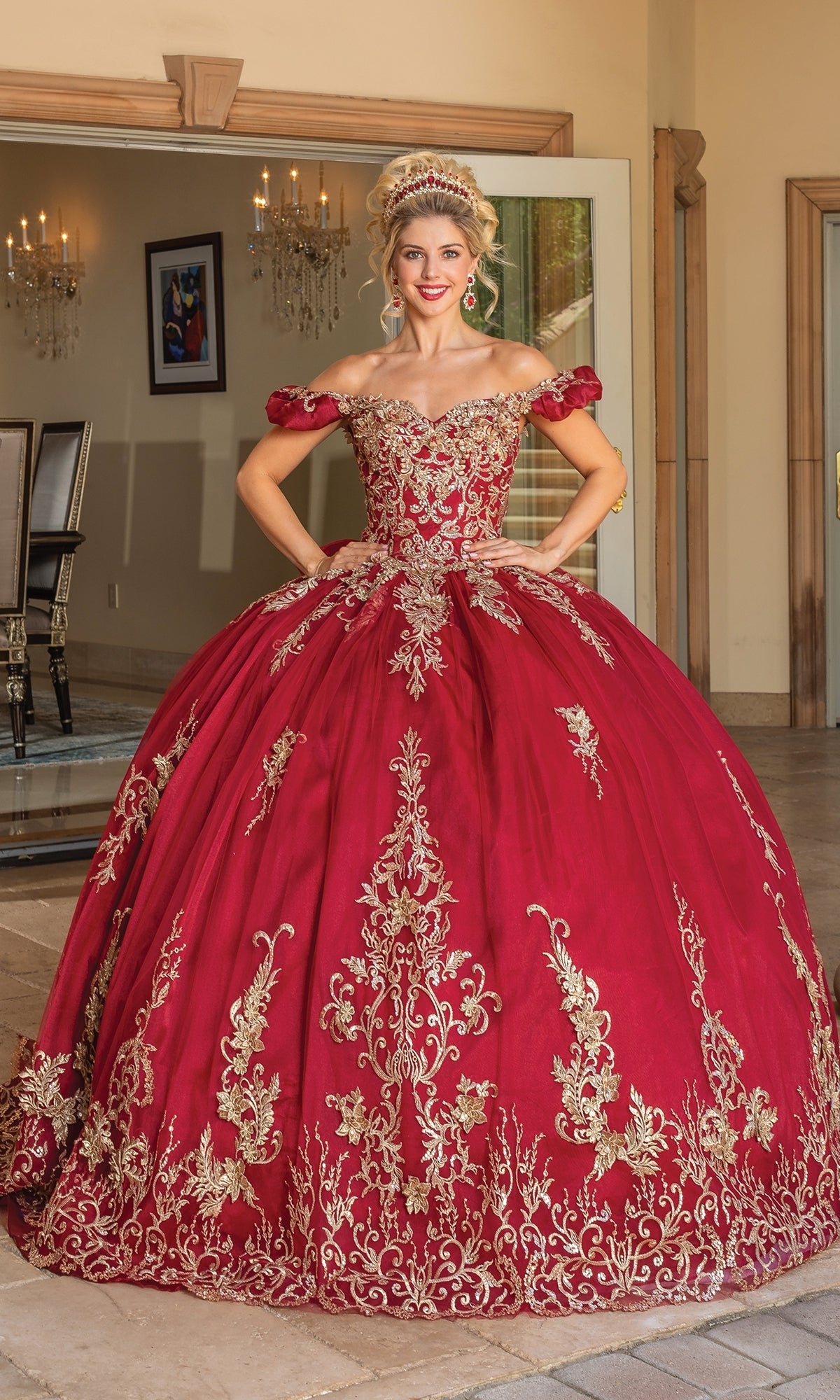 A woman stands indoors wearing Quinceanera Dress 1697 with Embroidery—an ornate, off-the-shoulder red gown featuring gold embroidery and a matching tiara.